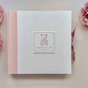 Ein handgebundenes Fotoalbum mit Letterpress Einband auf weißem Baumwollbüttenpapier mit Caromuster-Prägung und einer aufgeklebten quadratischen Vignette, die mit einem Teddy und Text in Rosa bedruckt ist