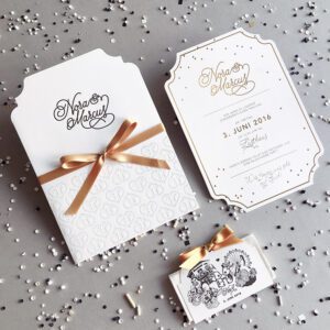 Hochzeitseinladung mit Letterpress Pockethülle mit farbloser Prägung und schwarzer Schrift bedruckt, über der Hülle ist ein goldener Band gebunden. In der Hülle steckt eine Karte mit Prägung und glänzend goldener Schrift. Eine taschentuchhülle ist auch sichtbar für die Freudentränen.