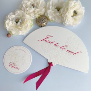 Letterpress Fächer und Glasuntersetzer auf leinenweißem Baumwollpapier mit floraler, farbloser Prägung und pinker Schriftzug und Satinband.