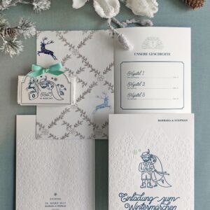 Einzigartige Letterpress Wintermärchen Hochzeitseinladung mit weißer Letterpress-Hülle in farbloser Prägung, die die Schneeflocken darstellt und eine winterliche Stimmung für die Alpenhochzeit erzeugt. Die Hülle zeigt auch in eisblauer Druckfarbe eine handgezeichnete Illustration des Brautpaares und eine geprägte Bergsilhouette, die das Brautpaar umgibt.